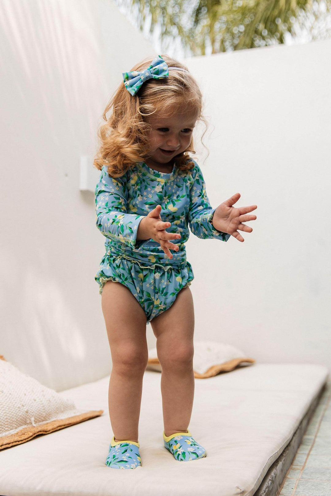 Bebezinha usando look de praia todo combinando na estampa Limões, está vestindo tapa fralda, camiseta UV com proteção, sapatinho neoprene e lacinho de cabelo