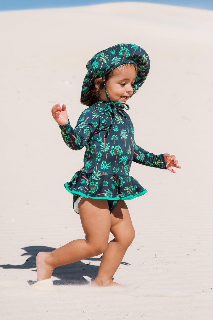 Bebe de 2 anos está vestindo maiozinho de praia infantil feminino com manga longa, babadinho, e zipper na frente, estampa coqueiros azul marinho com chapéuzinho combinando, da Lili Sampedro