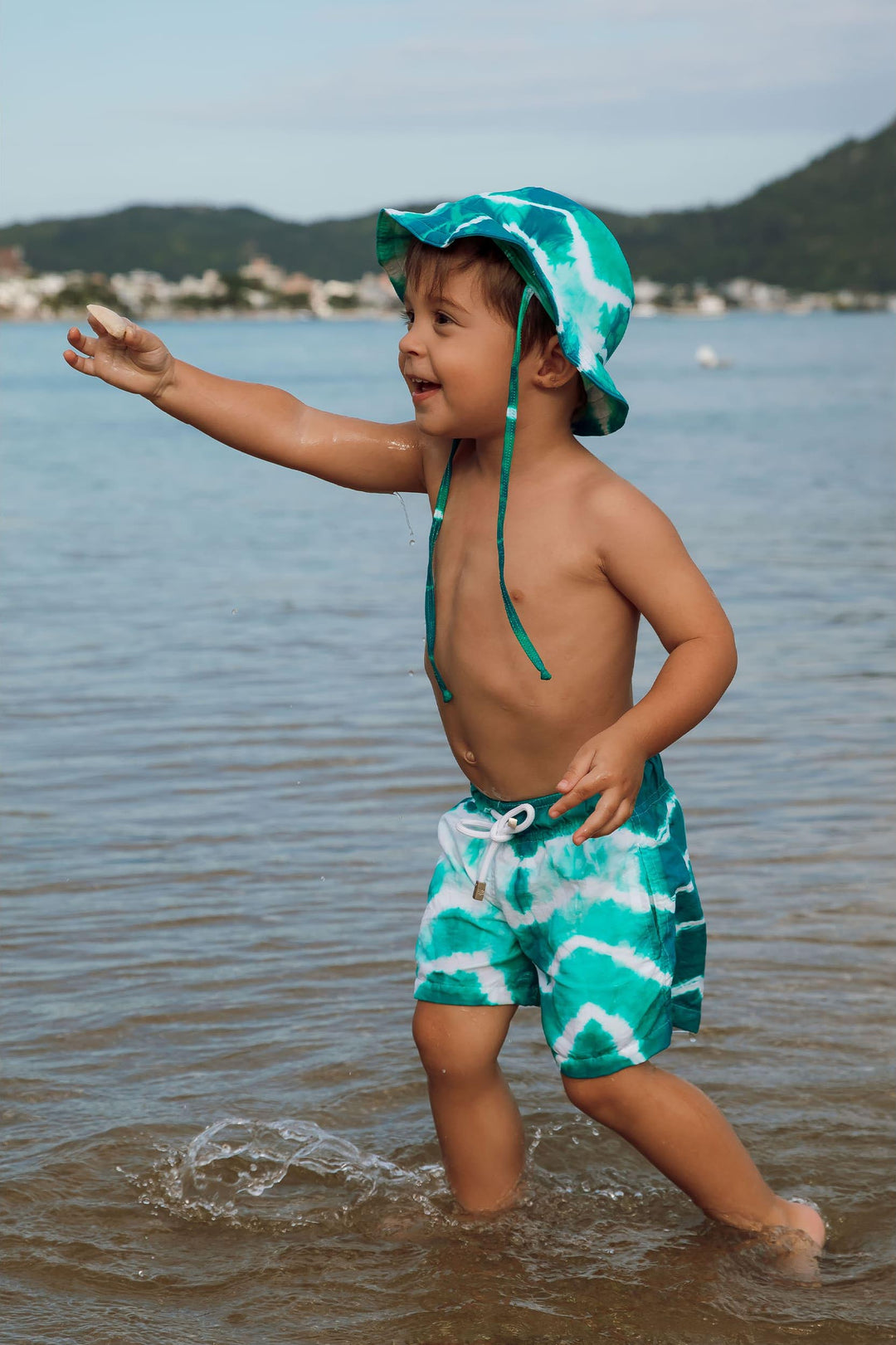 Menino de 2 anos vestindo short infantil de praia Tie Dye Verde com chapéuzinho infantil combinando da Lili Sampedro